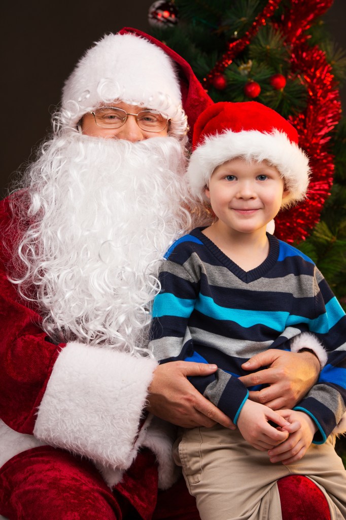 Funny little boy sitting on the lap of Santa Claus. Looking together into the camera and smiling.