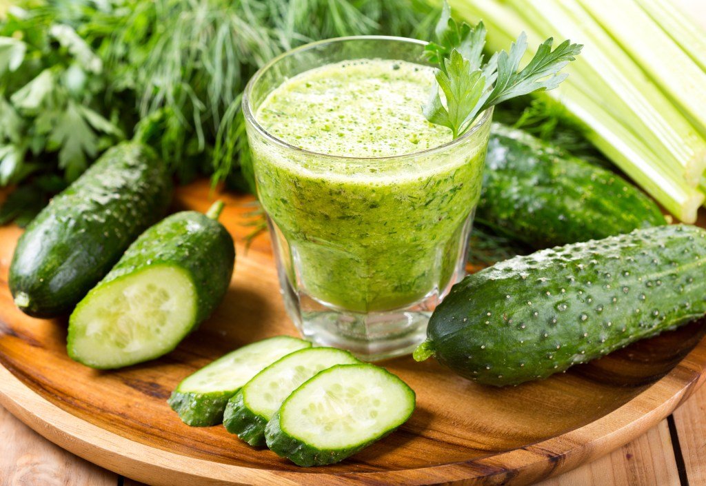 A glass of green juice surrounded by cucumbers with some celery and parsley in the background