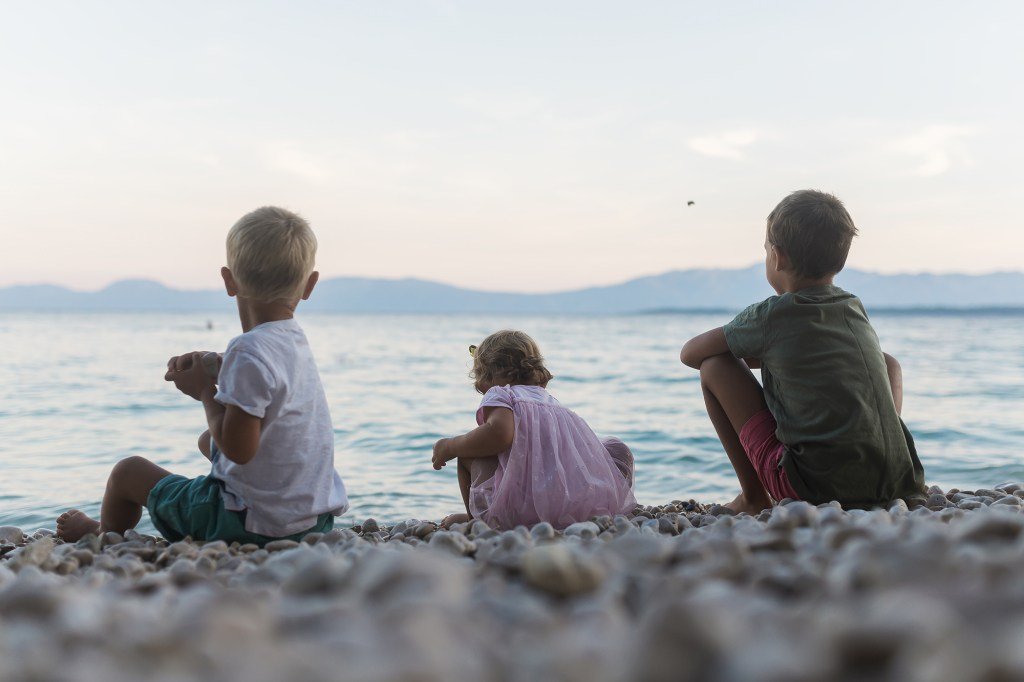 Three children, two sons and a girl, sitting on a pebble beach in the evening, throwing stones in the sea
