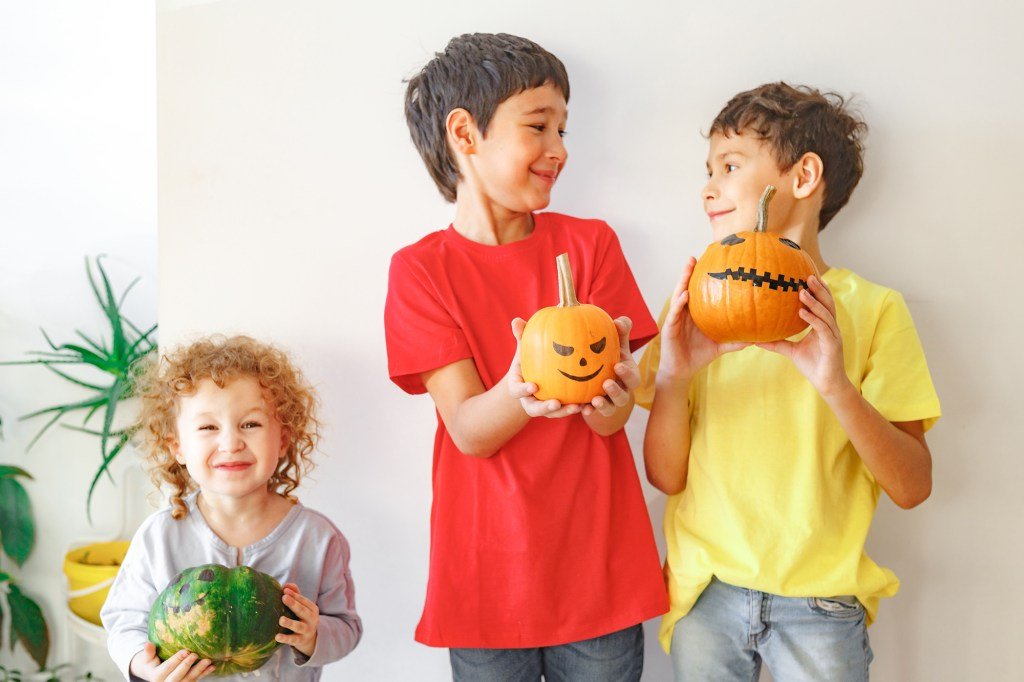 Three sisters and sisters standing against a white wall holding pumpkins carved with scary faces for Halloween