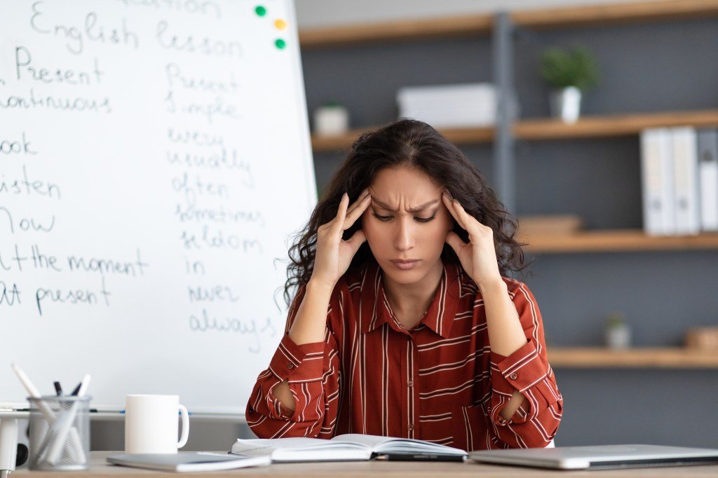 Young exhausted teacher touching the forehead and temples, suffering from a headache on her table near a white table